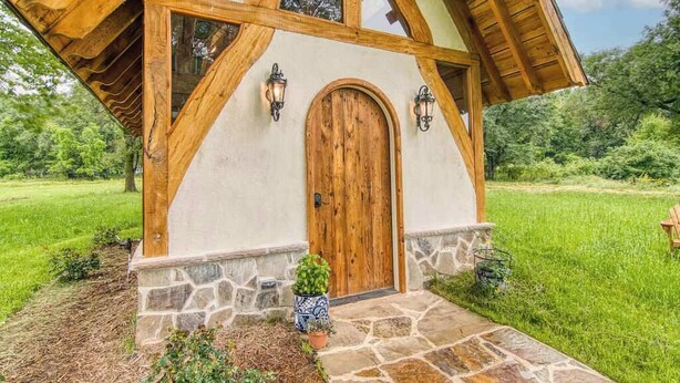 A rustic wooden cottage with a stone foundation, a wooden door, and two wall-mounted lanterns. - Experience a Regenerative Farm in a New Tiny Home (Nixa)
