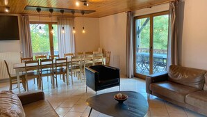 Ein geräumiges Wohnzimmer mit Holzbalkendecke, einem Essbereich mit Holzsesseln und einem Tisch, einem Ledersofa, einem schwarzen Couchtisch mit einer Schüssel und großen Fenstern mit Vorhängen.