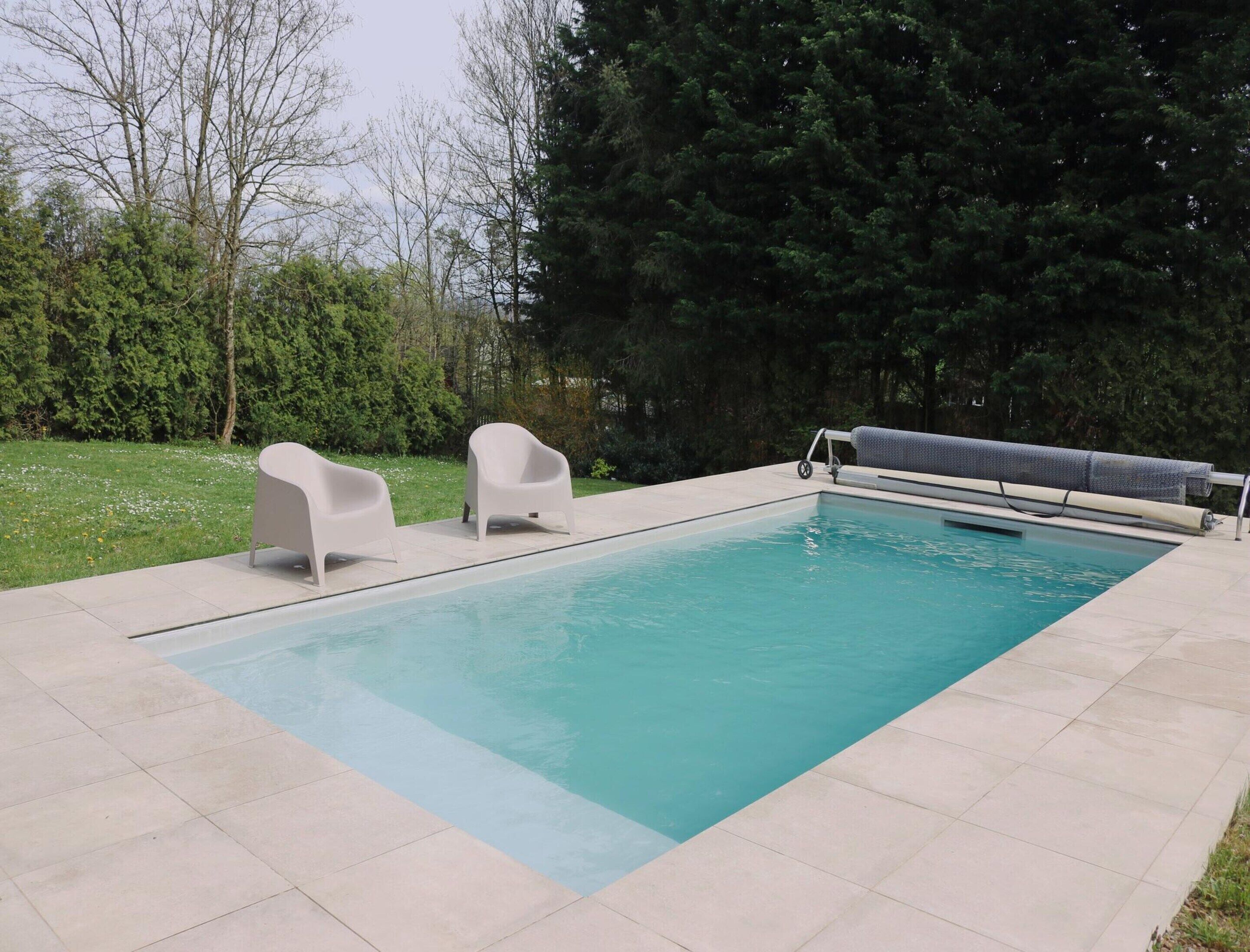 A rectangular swimming pool with a tiled deck, two white lounge chairs, and a covered bench.