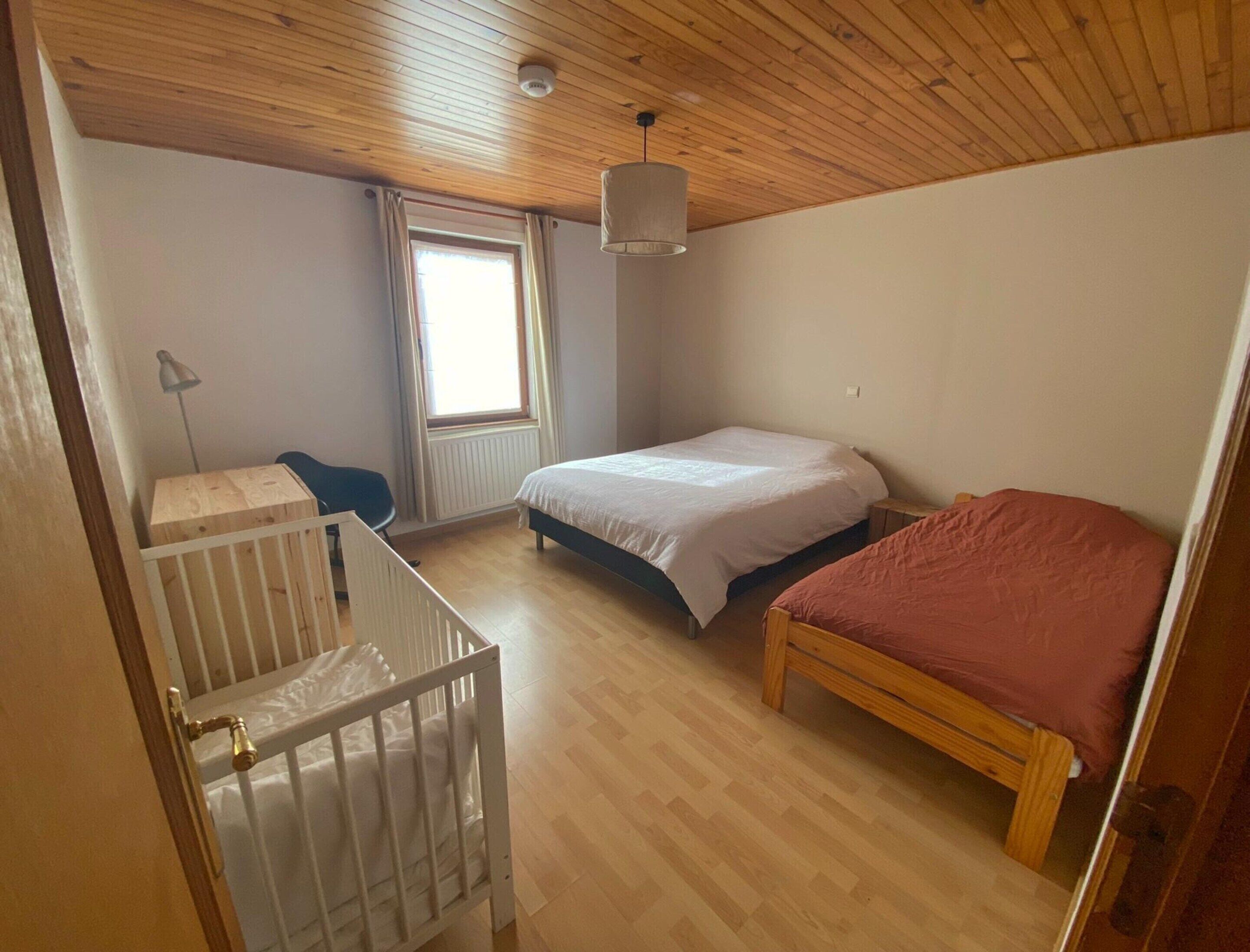 A bedroom with a wooden ceiling, a window with curtains, a bed with a white and red bedspread, and a crib with a white mattress.