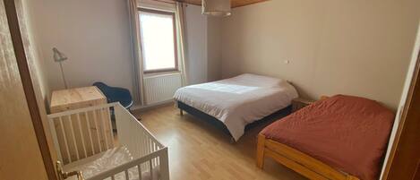 Um quarto com teto de madeira, janela com cortinas, cama com colcha branca e vermelha, e berço com colchão branco.