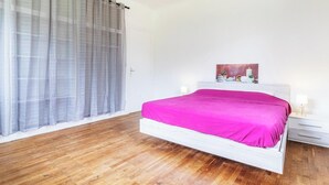 A bedroom with a wooden floor, a bed with a pink bedspread, a white headboard, a bedside table with a lamp, and a window with sheer curtains.