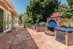 A patio with a brick barbecue area, a glass-enclosed room, and lush greenery.