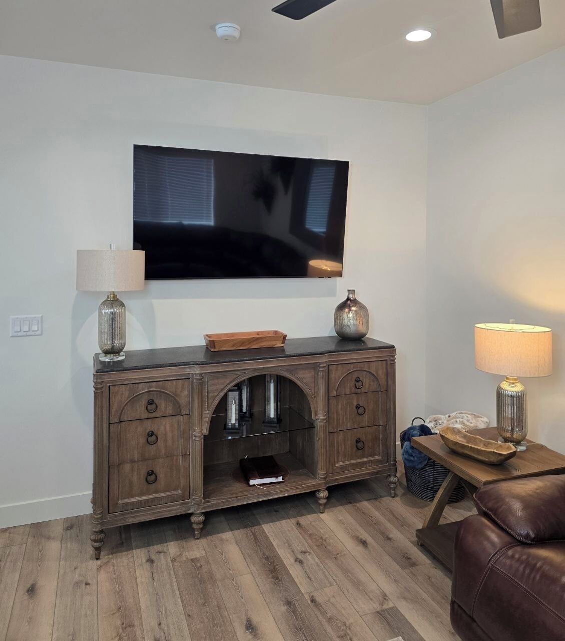 Una sala de estar con un televisor de pantalla plana montado en la pared, un mueble de madera con cajones, un sofá de cuero y una mesita de noche con una lámpara.
