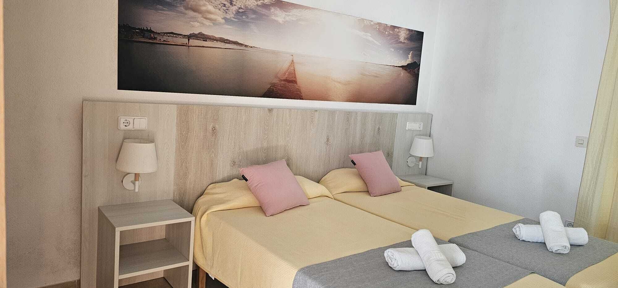 Una habitación de hotel con una cama, dos mesitas de noche, una lámpara y un cuadro con un atardecer sobre un puente.