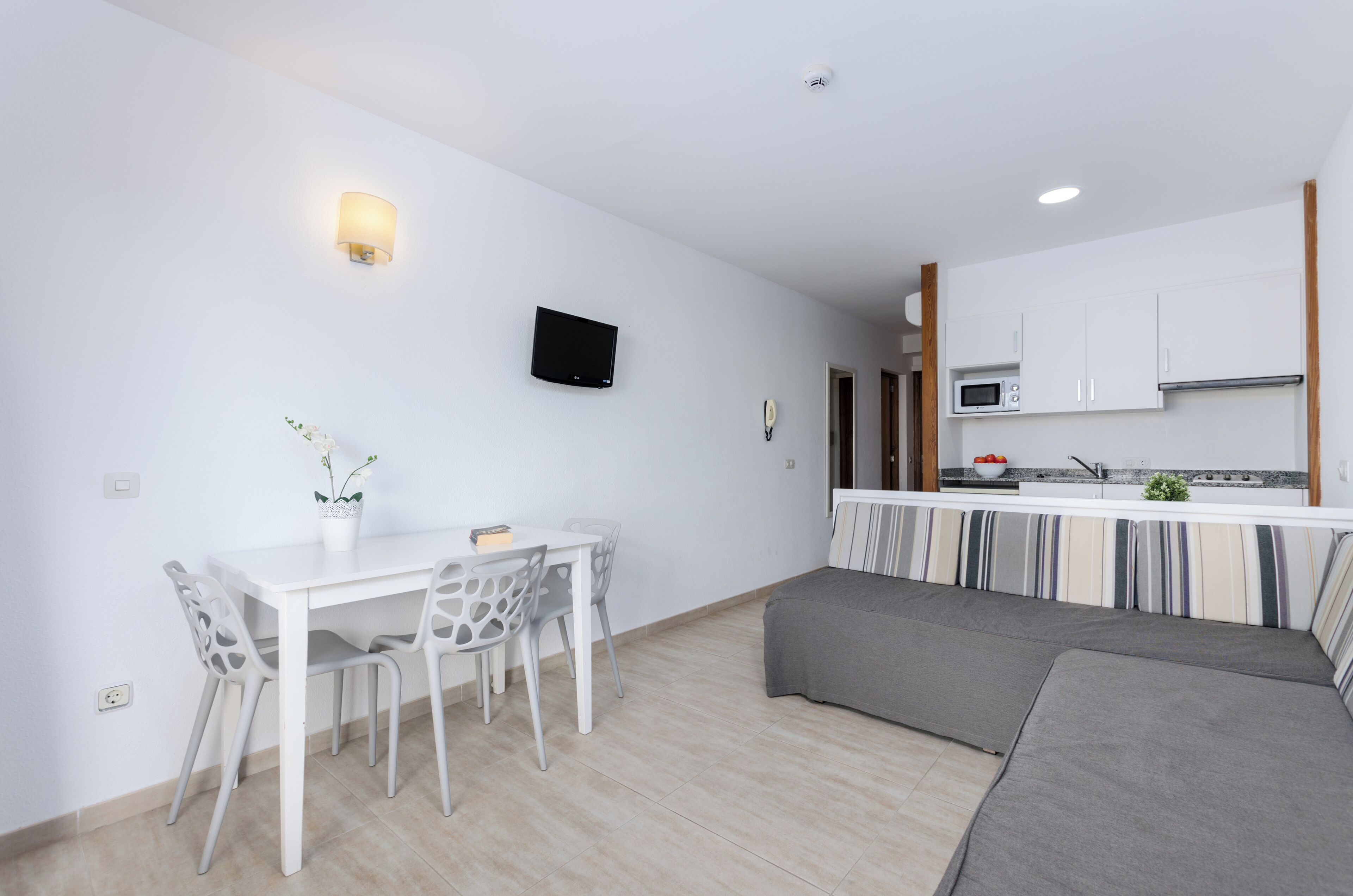 A modern kitchen with white cabinets, a microwave, and a sink. There is a dining area with a white table and chairs, and a grey sofa.
