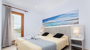 A hotel room with a bed, a nightstand, a painting of a beach, and a view of the sea through the window. - Apartamentos Venecia  (Alcúdia)