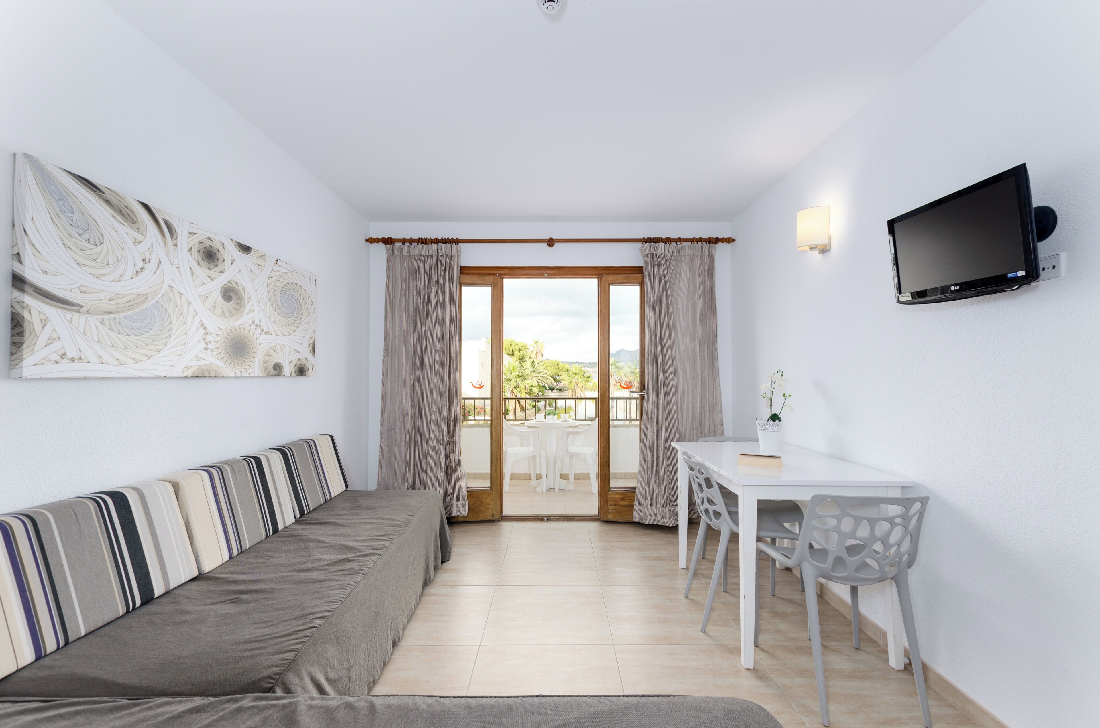 Una habitación moderna con sofá, una mesa de comedor con sillas, televisor montado en la pared y un gran ventanal con cortinas.
