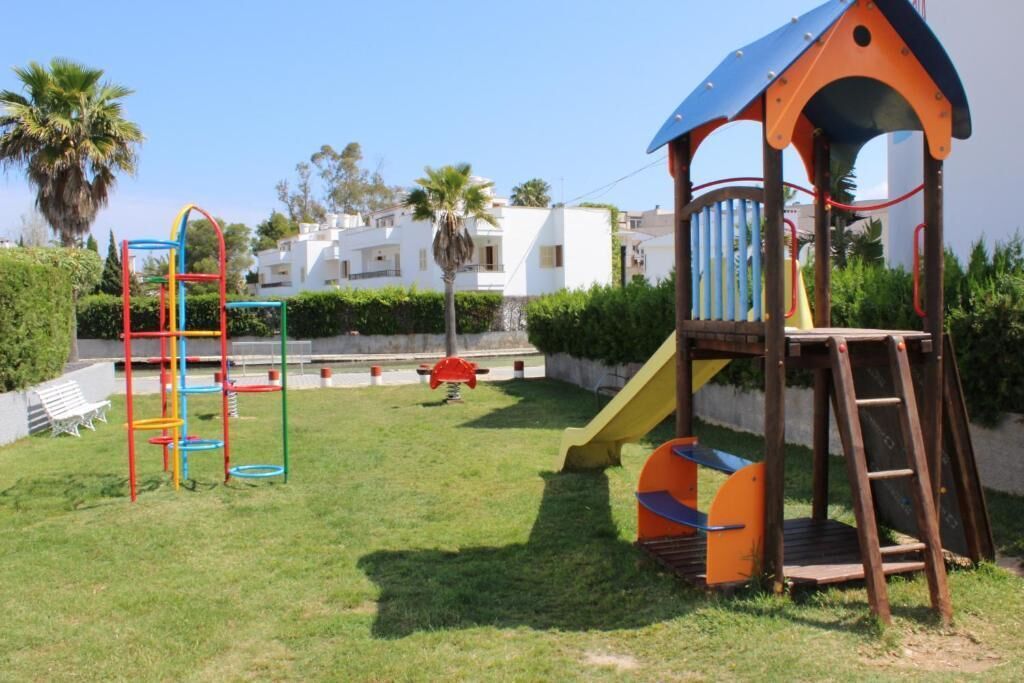 A playground with a slide and climbing structures, surrounded by a grassy area and a white building in the background.