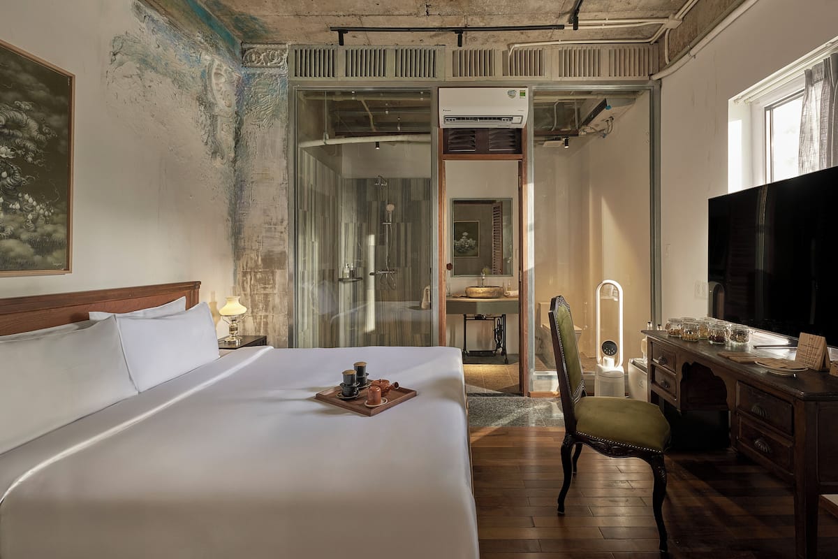 A hotel room with a bed, a desk with a TV, a chair, a bathroom with a shower, and a painting on the wall.