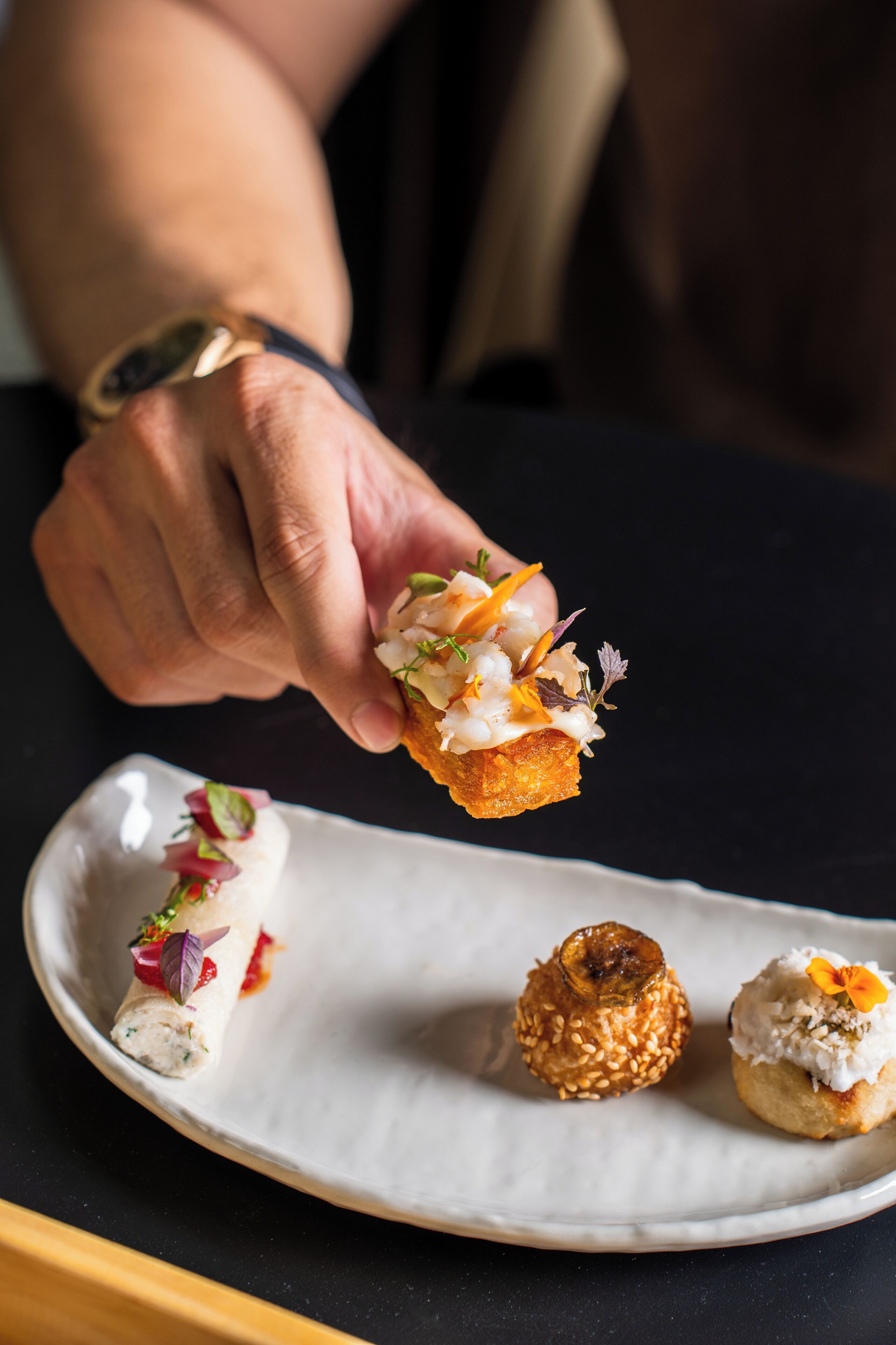 A hand holding a food item over a plate with three other food items.