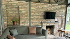 A cozy living area with a brick fireplace, a sofa with cushions, a television mounted above the fireplace, and a small table with a plant. - Junior Garden Villa (Sapanca)