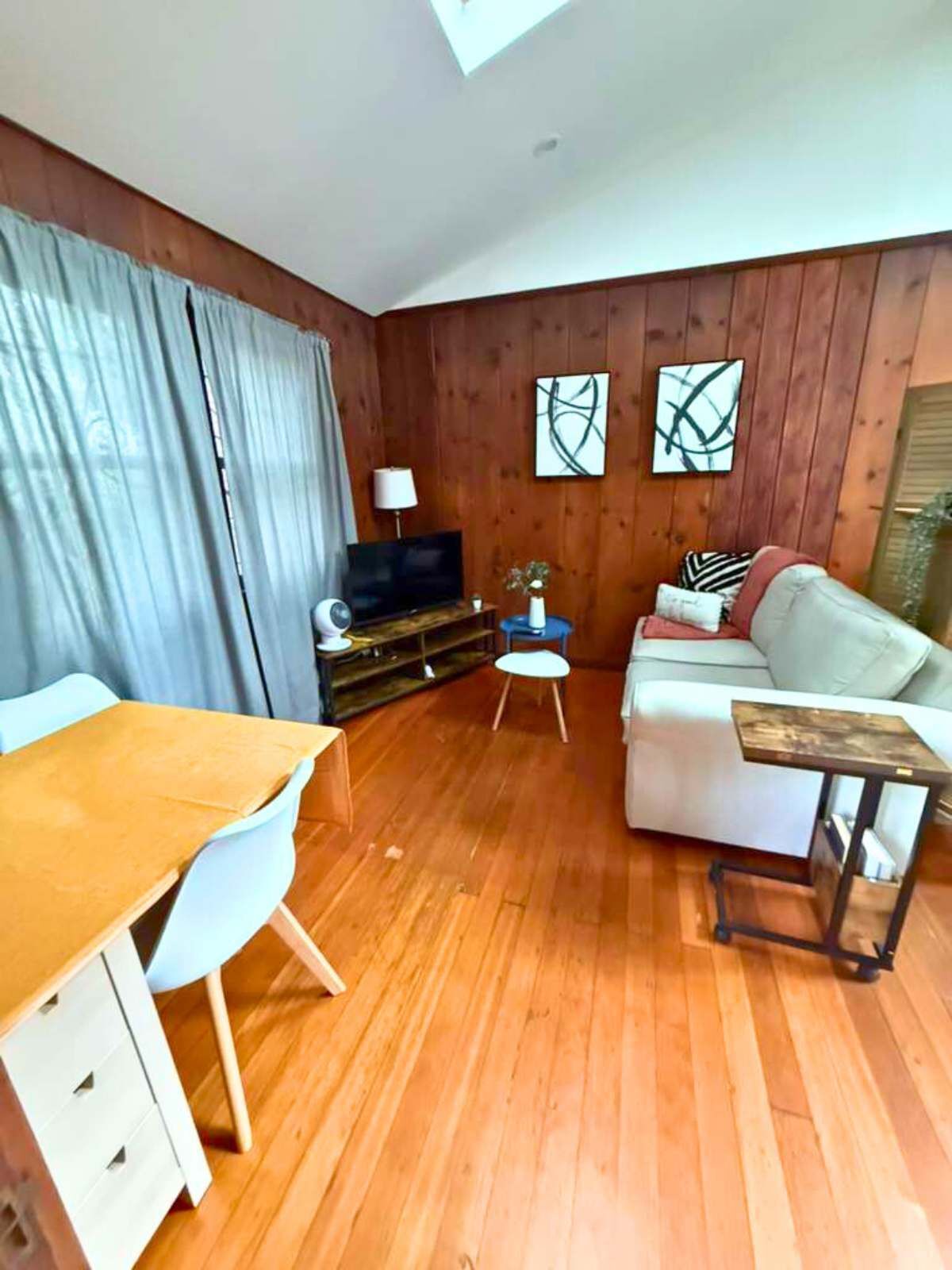 Un salon doté d’un plancher en bois, d’un canapé blanc, d’une table basse en bois, d’un téléviseur posé sur un meuble en bois et d’une table à manger avec des chaises.