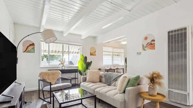 A modern living room with a sofa, coffee table, and TV. There are windows with blinds, a plant, and artwork on the walls.