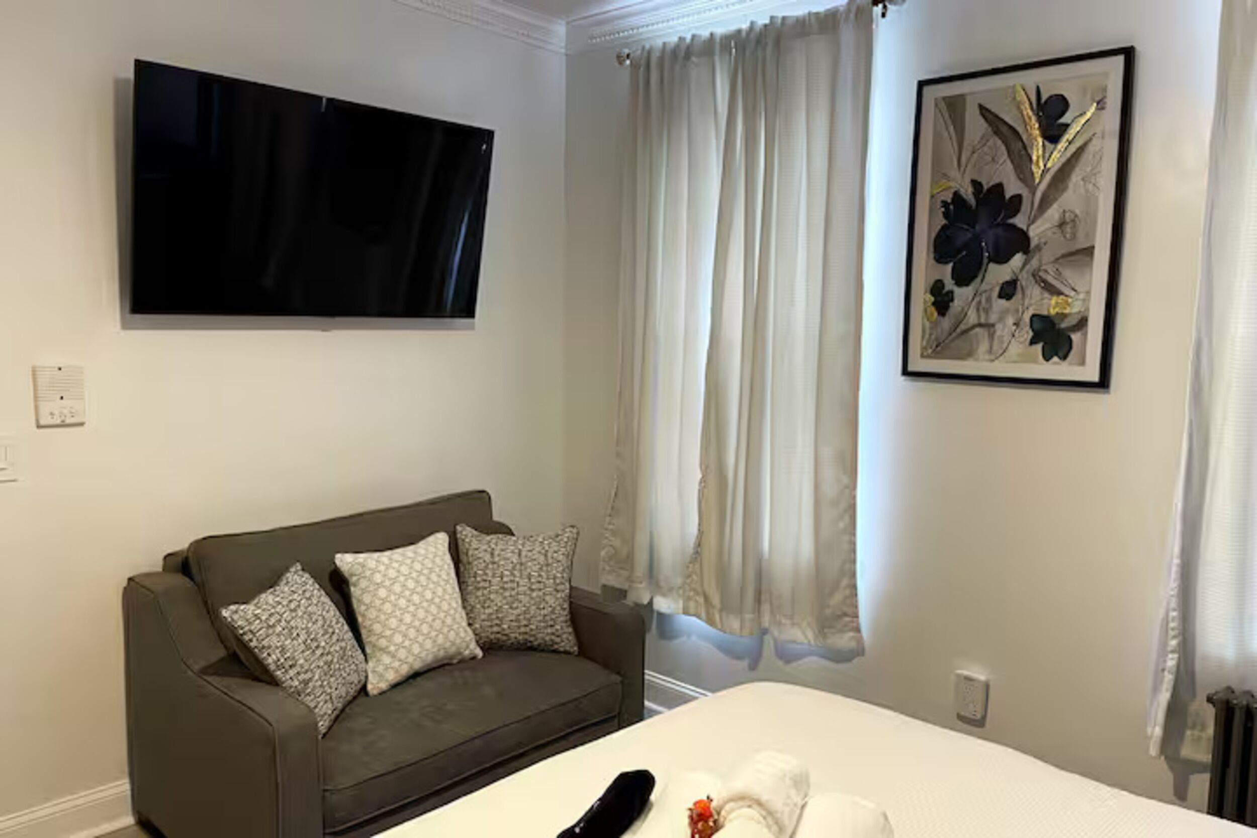 A hotel room with a bed, a grey sofa, a flat-screen TV, and a framed floral artwork.