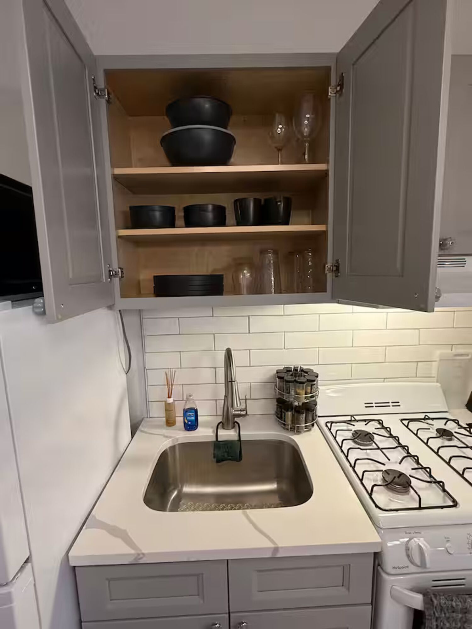 A kitchen with a white countertop, a stainless steel sink, a stove, and open cabinets with dishes and glasses.
