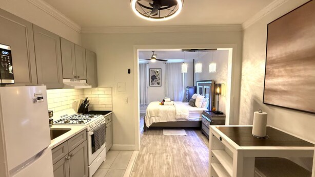 A modern kitchen with white cabinets, a gas stove, and a refrigerator. There is a hallway leading to a bedroom with a bed and a nightstand. - WORLD CUP + minutes to NYC (Union City)