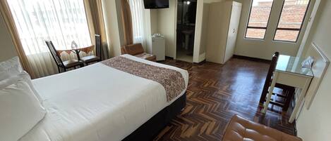 Habitación de hotel con una cama grande, un escritorio, una silla, una mesita, un televisor y un baño visible a través de una puerta abierta.
