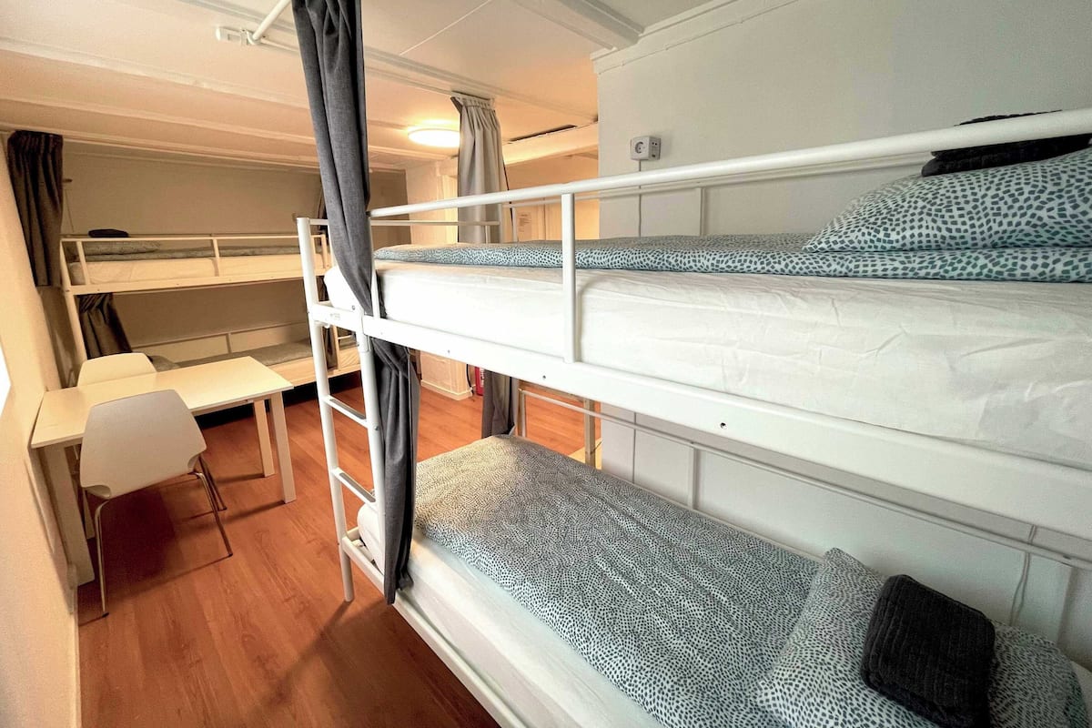 Une chambre avec un lit superposé, un bureau et une chaise, une petite table et une fenêtre avec des rideaux.