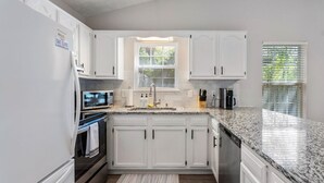 A kitchen with white cabinets, a granite countertop, a microwave, a coffee maker, and a window with blinds. - Golf Cart, Bikes, firepit, & More! (Oak Island)