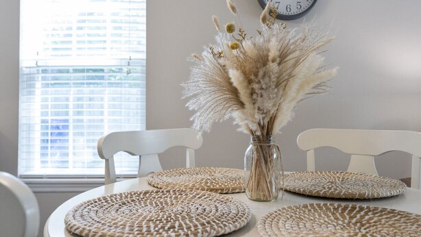 A dining table with woven placemats, a vase with dried pampas grass, and a wall clock. - Golf Cart, Bikes, firepit, & More! (Oak Island)