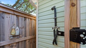 A wooden fence with a metal rack holding cleaning supplies, a black hose attached to a wall-mounted faucet, and a black electrical box. - Golf Cart, Bikes, firepit, & More! (Oak Island)