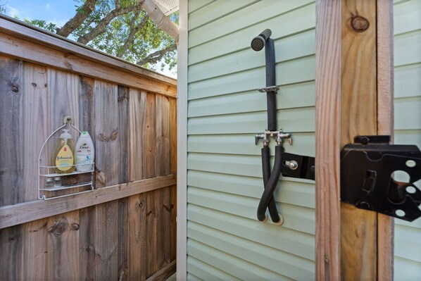A wooden fence with a metal rack holding cleaning supplies, a black hose attached to a wall-mounted faucet, and a black electrical box. - Golf Cart, Bikes, firepit, & More! (Oak Island)