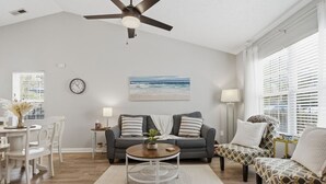 A living room with a gray sofa, a coffee table, and a patterned armchair. There is a painting on the wall, a clock, and a window with blinds. - Golf Cart, Bikes, firepit, & More! (Oak Island)