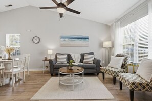 A living room with a gray sofa, a coffee table, and a patterned armchair. There is a painting on the wall, a clock, and a window with blinds.
