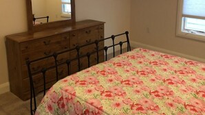 Une chambre avec une commode en bois, un lit recouvert d’une couvre-lit à motifs floraux, un miroir au-dessus de la commode et une fenêtre avec des stores.