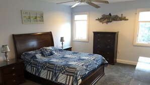Une chambre à coucher comprenant un lit, une table de chevet, une commode, un ventilateur de plafond et une fenêtre.