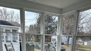 Una mecedora blanca en una sala de estar con ventanales amplios, piso de madera y vista a una calle residencial.