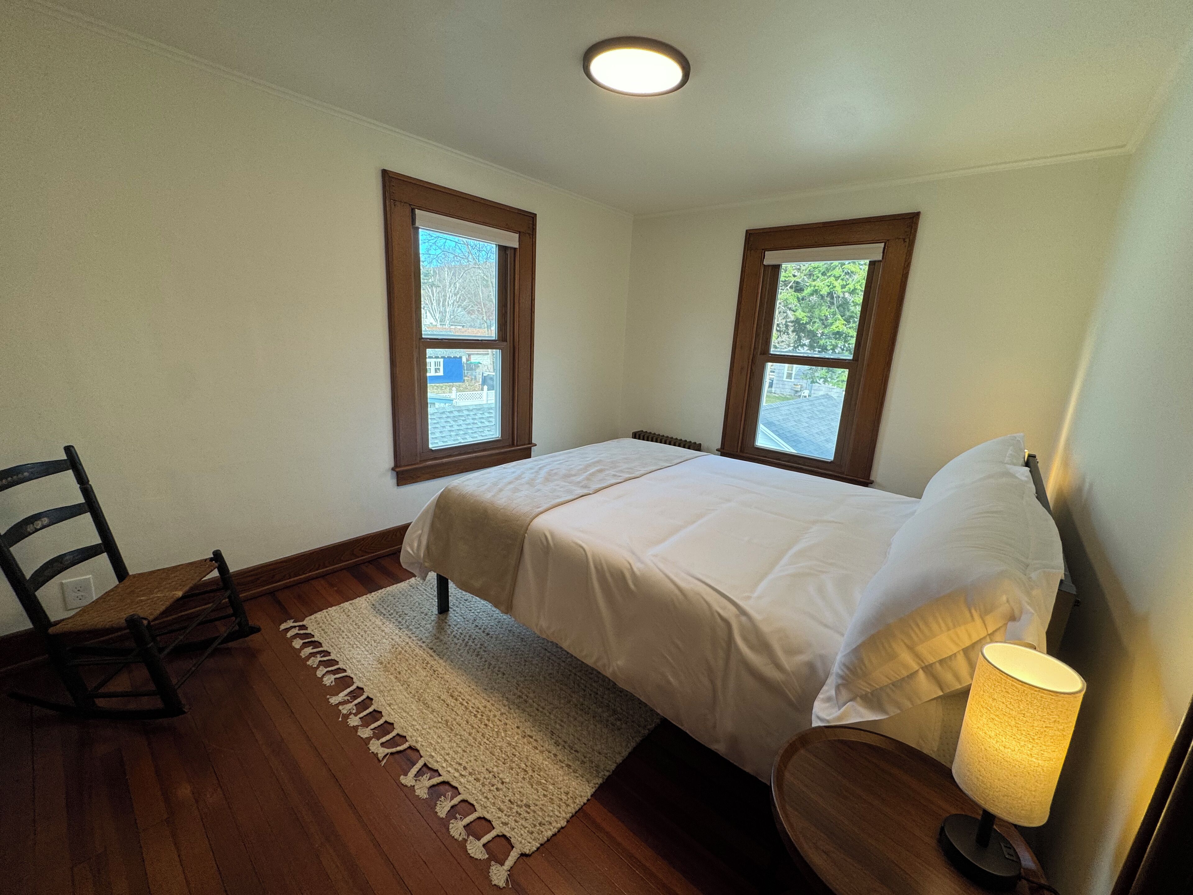 A bedroom with a bed, two windows, a chair, a lamp, and a rug.