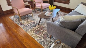 Un salón con suelo de madera, un sofá gris, dos sillones rosa, una mesa de centro con un libro y flores, y una alfombra estampada.