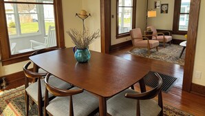 Un comedor con una mesa y sillas de madera, un jarrón con flores secas y vistas al exterior a través de amplias ventanas.
