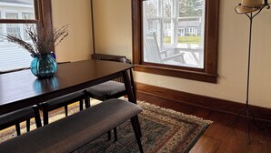 Un comedor con mesa de madera oscura, bancos y un jarrón con flores secas. Amplias ventanas de marco blanco ofrecen vistas a una casa y un jardín.