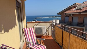 A balcony with two striped lounge chairs, an air conditioning unit, and a view of the sea and buildings.