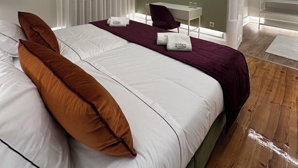 Une chambre d’hôtel moderne avec un grand lit, un bureau et une chaise. On y trouve deux lampes fixées au mur et une fenêtre avec des stores.