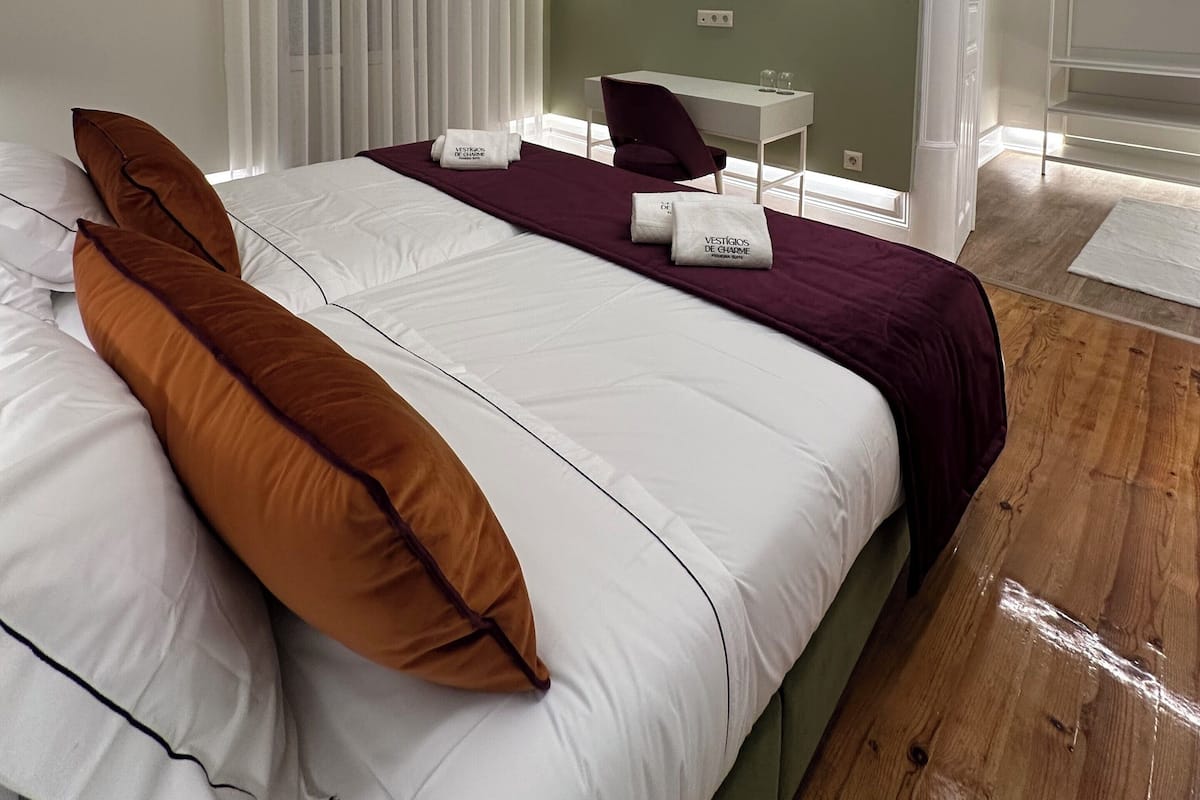 Une chambre d’hôtel moderne avec un grand lit, un bureau et une chaise. On y trouve deux lampes fixées au mur et une fenêtre avec des stores.