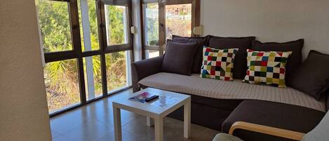 A living room with a sofa, a coffee table, and large windows offering a view of greenery and mountains.