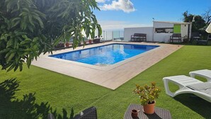 Una piscina rectangular rodeada de césped artificial, con una pequeña zona de patio y un edificio blanco al fondo.