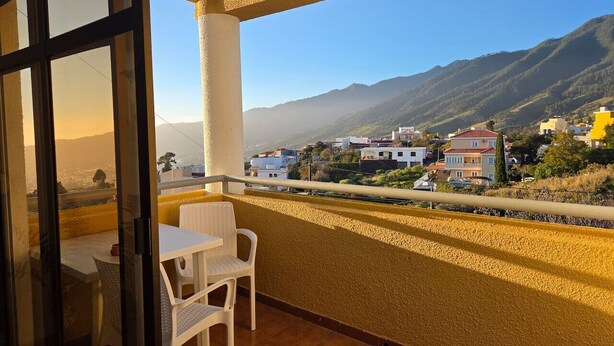 Un balcón con mesa y sillas, con vistas a una colina con edificios y montañas.