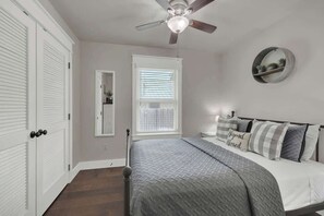 A bedroom with a bed, a ceiling fan, a window with blinds, and a wall-mounted shelf with plants.