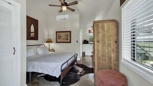 A bedroom with a bed, a ceiling fan, a wooden headboard, a window with blinds, and a small stool. - Casa Salvia Blanca + Rojo & Tejas Combo! (Fredericksburg)
