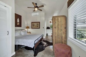 A bedroom with a bed, a ceiling fan, a wooden headboard, a window with blinds, and a small stool.