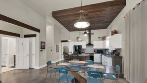 A spacious kitchen with modern appliances, a dining area with a large table and blue chairs, and a high ceiling with exposed beams. - Casa Salvia Blanca + Rojo & Tejas Combo! (Fredericksburg)