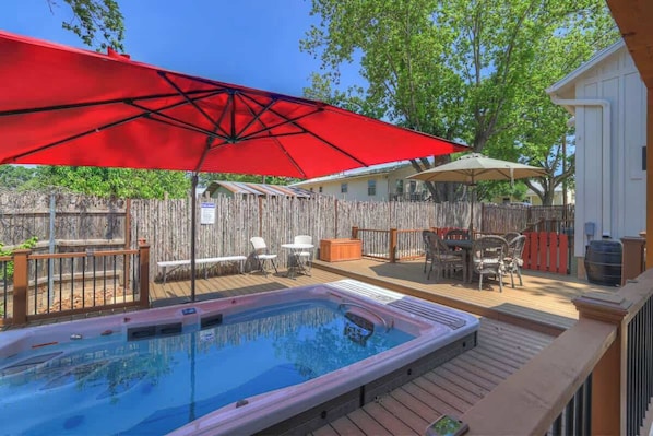 Un patio trasero con jacuzzi, terraza de madera, mobiliario exterior y un paraguas rojo.