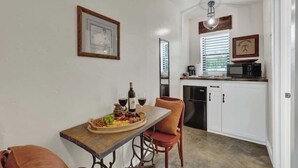 A compact kitchen with white cabinets, a microwave, and a coffee maker. A dining table with a fruit platter and wine glasses is set against the wall. - Casa Salvia Blanca + Rojo & Tejas Combo! (Fredericksburg)