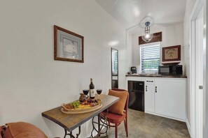 Uma cozinha compacta com armários brancos, micro-ondas e cafeteira. Uma mesa de jantar com uma travessa de frutas e copos de vinho está posicionada junto à parede.