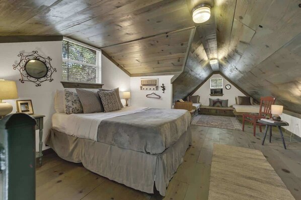 Ein gemütliches Schlafzimmer im Dachgeschoss mit einem großen Bett, einem Schreibtisch und einem Stuhl. Ein Fenster sorgt für natürliches Licht und bietet Blick auf Bäume.